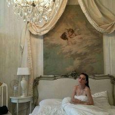 a woman sitting on top of a white bed in a bedroom next to a chandelier