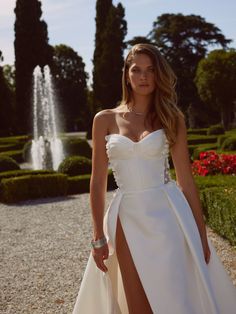 a woman in a white wedding dress posing for the camera with a fountain behind her