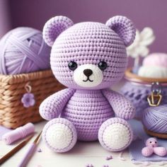 a crocheted purple teddy bear sitting next to yarn and knitting needles on a table