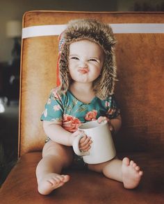 a baby with a furry hat sitting on a chair holding a coffee cup and smiling at the camera