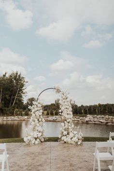 an outdoor ceremony setup with white chairs and flowers on the aisle, overlooking a pond