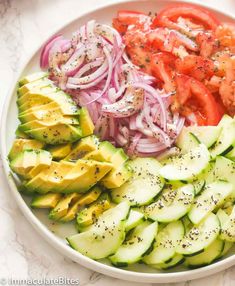 a white plate topped with cucumber, tomatoes and onions next to sliced avocado