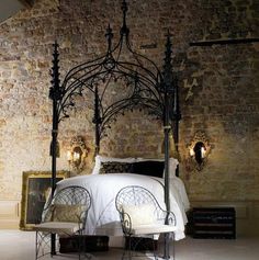 an iron bed frame with white sheets and pillows in a brick walled bedroom, along with two chairs