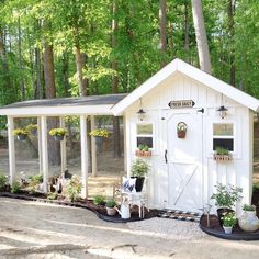 a small white shed sitting in the middle of a forest with potted plants on it