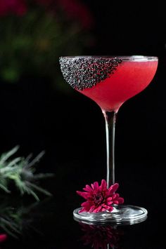 a red drink in a wine glass with black glitter on the rim next to a pink flower