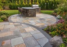 an outdoor kitchen and dining area in a backyard with stone pavers, flowers and trees