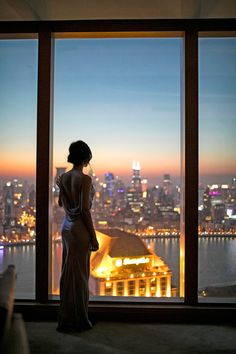 a woman standing in front of a window looking out at the city lights and water