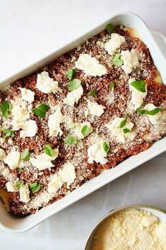 a casserole dish with cheese and herbs on top, next to a bowl of powdered sugar