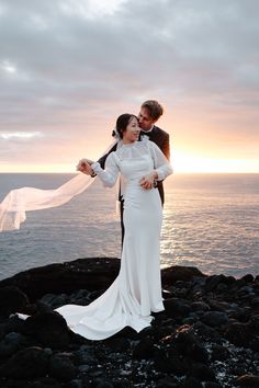 a bride and groom standing on rocks by the ocean at sunset with their veil blowing in the wind