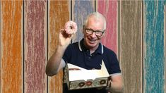 an older man holding up a doughnut in front of a colorful wall with stripes