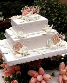 a three tiered wedding cake with sea shells and corals on the top is surrounded by greenery