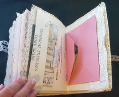 an open book with pink paper and white lace on the pages is being held by a hand