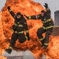 two firemen jumping in the air with their arms up and legs apart, surrounded by flames