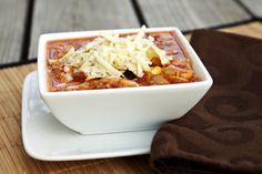 a white bowl filled with chili and cheese on top of a wooden table next to a brown napkin