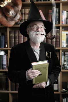 an older man in a black suit and hat holding a blue book while standing in front of a bookshelf