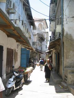 two people walking down an alley way with buildings in the background and one person on a scooter