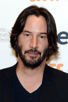 a man with long hair and beard wearing a black suit standing in front of a white wall