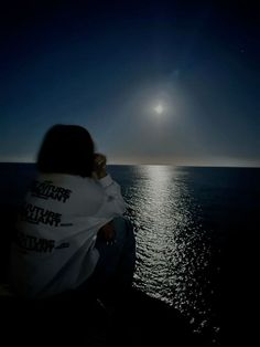 a person sitting on the edge of a cliff looking out at the ocean under a full moon