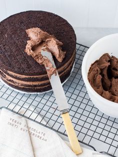 a chocolate cake on a cooling rack with a knife and bowl of frosting next to it