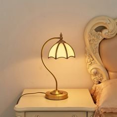 a lamp on a night stand next to a white dresser with a bed in the background