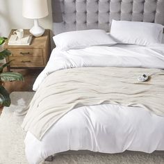 a bed with white linens and pillows in a bedroom next to a plant on the floor