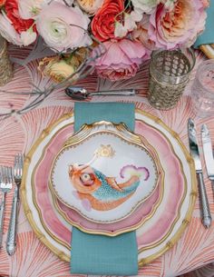 a pink and gold plate with a mermaid design on it, surrounded by silverware