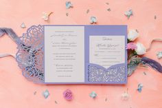 an open purple and white wedding card on a pink surface with flowers in the background