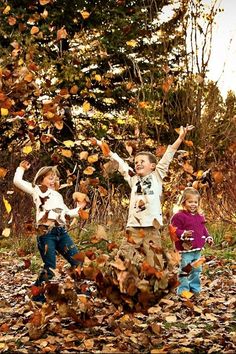 three children throwing leaves in the air on top of a leaf covered ground with trees behind them