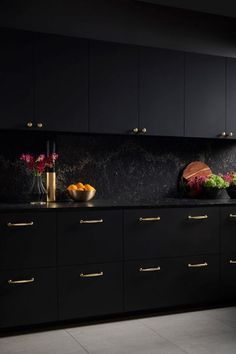 a kitchen with black cabinets and gold pulls on the countertop, along with bowls of fruit and vegetables