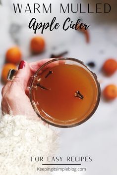 a person holding a glass filled with liquid and oranges in the background text reads warm mulled apple cider