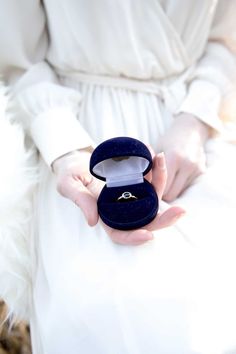 a woman in white dress holding an open ring box with a blue velvet cover on it