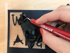 a hand holding a red marker over a piece of black paper on top of a wooden table