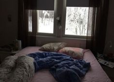 an unmade bed with blankets and pillows on it in front of two large windows