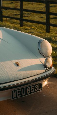 an old white car parked in front of a wooden fence with grass and flowers behind it