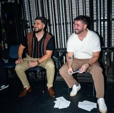 two men sitting next to each other on chairs in front of a wall with bars