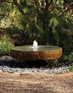 a water fountain in the middle of a garden with rocks and gravel around it, surrounded by greenery