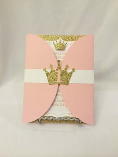 a pink and gold card with a crown on it's front, sitting on a white background