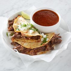 a white plate topped with beef and tortillas next to a bowl of salsa