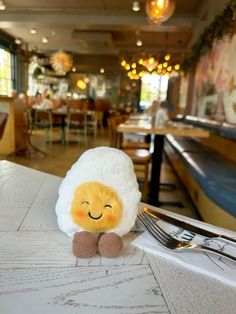 a stuffed sheep sitting on top of a table next to a fork and knife with a restaurant in the background