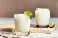 two glasses filled with ice and limes on top of a cutting board next to each other