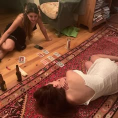 two women laying on the floor playing cards with beer bottles and papers scattered around them