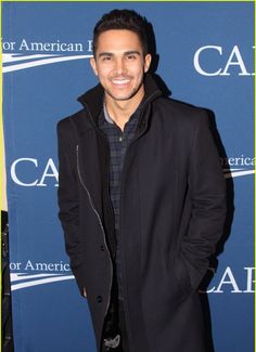 a man standing in front of a blue wall wearing a black coat and plaid shirt