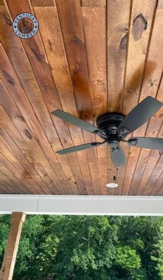 a ceiling fan mounted to the side of a wooden porch with trees in the background