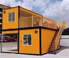 a yellow shipping container with stairs leading up to the second floor and another building in the background