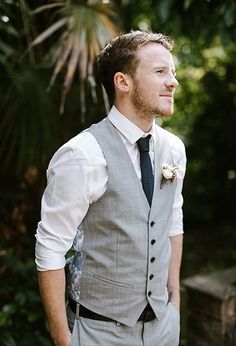 a man wearing a suit and tie standing in front of some trees with his hands in his pockets