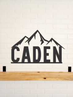 a metal sign that says caden with mountains in the background on a wooden shelf