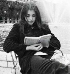 a young woman sitting on a bench reading a book in front of a water fountain