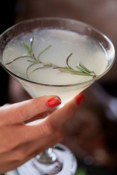 a woman holding a martini glass with white liquid and rosemary sprigs on it