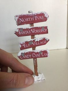 a hand holding a small wooden sign that says north pole santa's workshop, el village candy cane lane