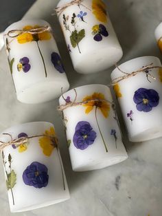 six white candles with purple and yellow flowers on them sitting on a marble counter top